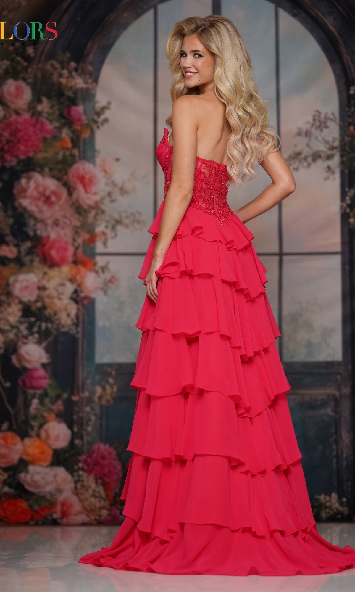 A woman with long blonde hair smiles in front of a floral, arched window backdrop wearing the Colors Dress 3934, a formal strapless long ruffled prom dress.